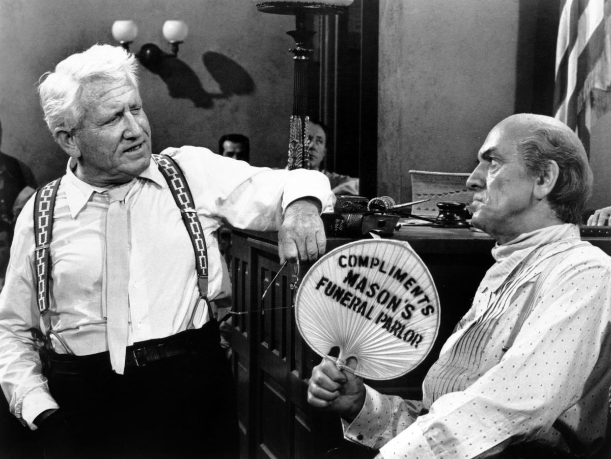 Spencer Tracy and Fredric March in Inherit the Wind. March is sitting in the witness box of a courtroom, fanning himself with a fan that says “Compliments of Mason’s Funeral Parlor,” as Tracy cross examines him. Both men are in their shirtsleeves. 