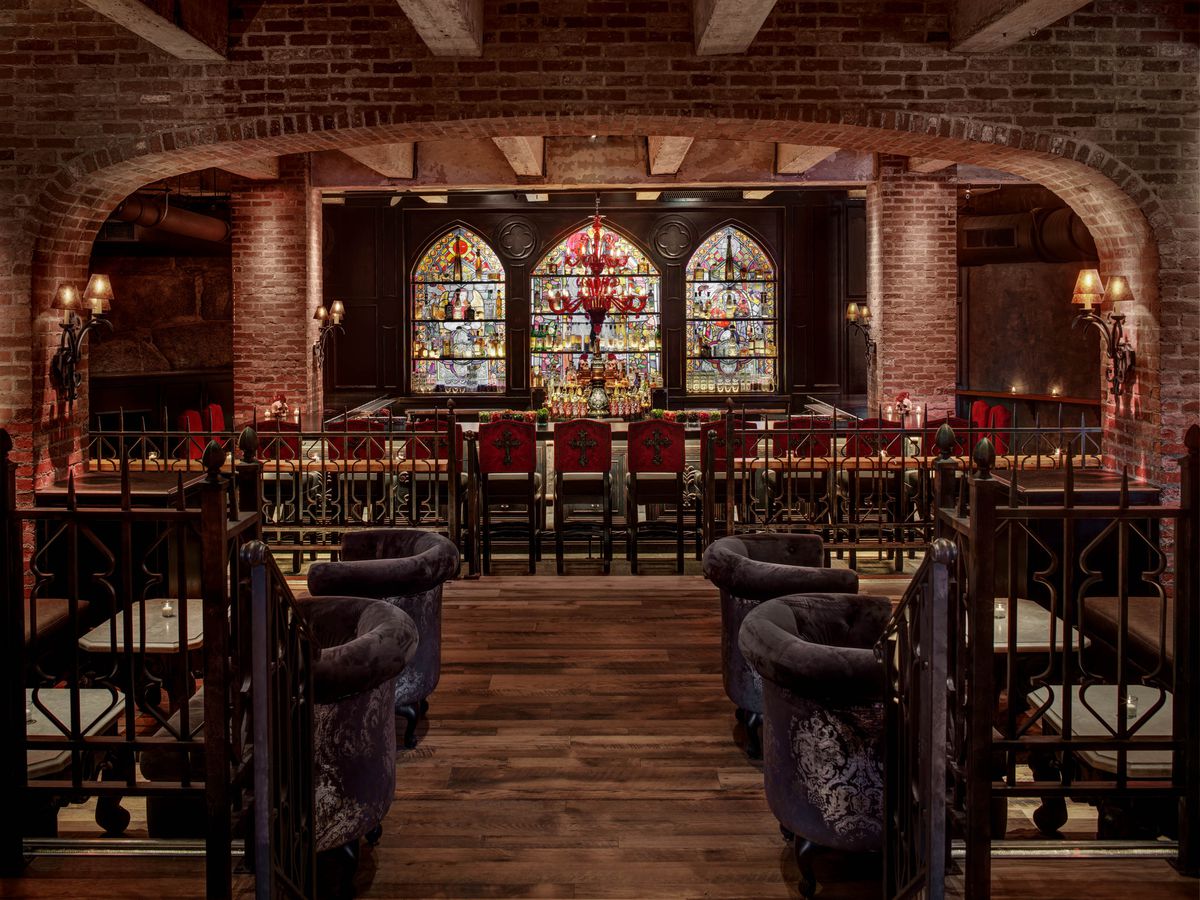 This view of a restaurant interior features a brick-lined, cavernous space. Red seats with crosses on their backs sit in front of a bar, which has three distinctive stained glass panels behind it.