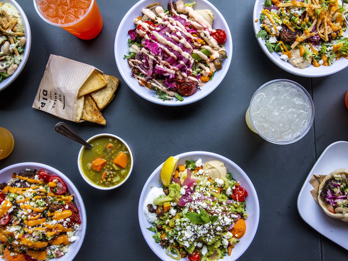 A spread of salads and grain bowls from Cava, arranged on a dark grey background.