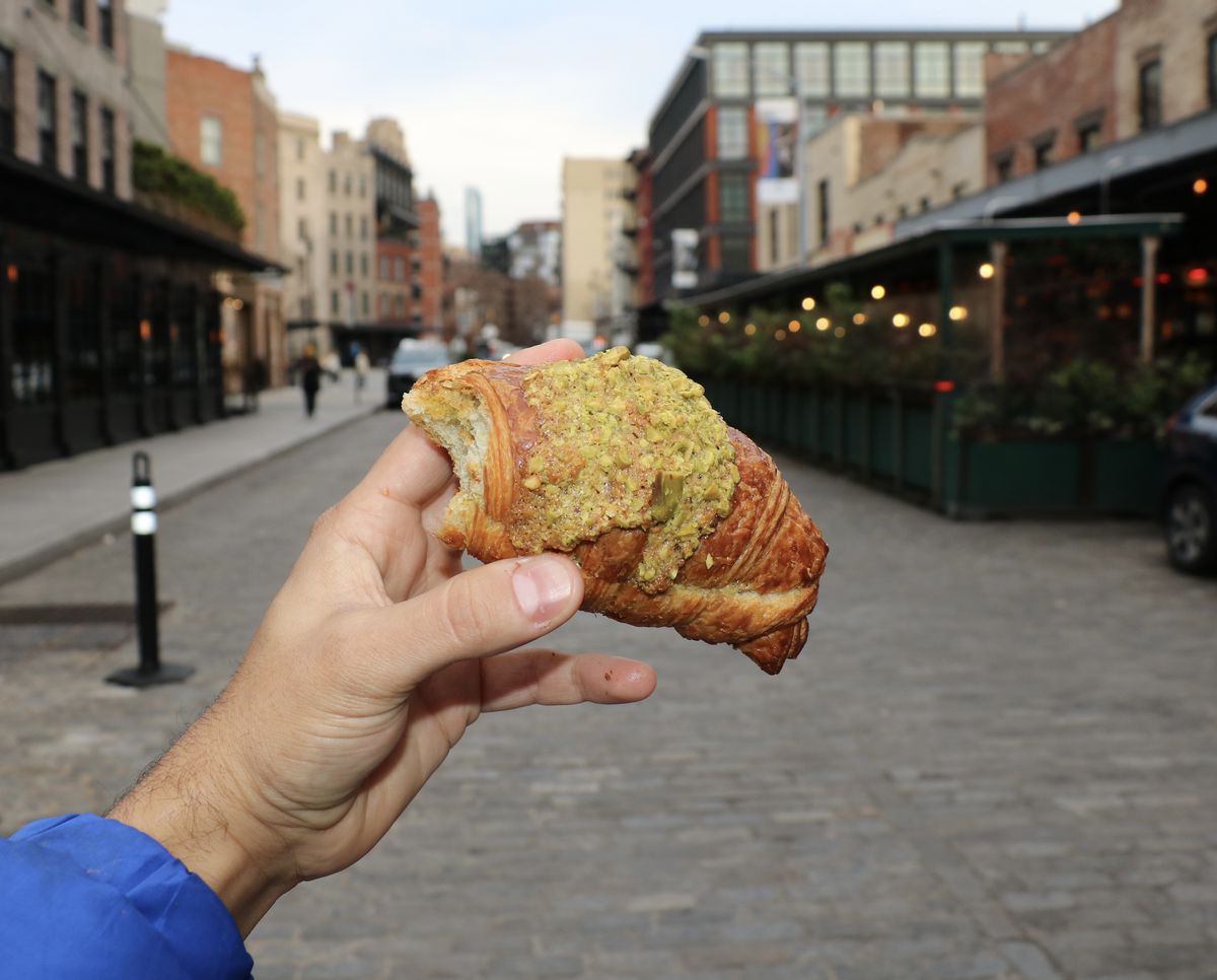 A hand holds up a pistachio croissant for a photograph on a cobblestone street.
