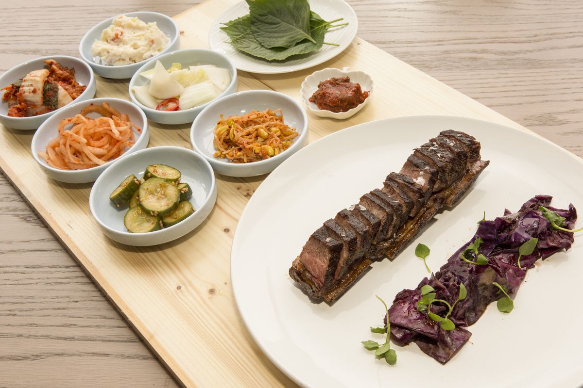 A wooden board holds a round white plate that contains sliced kalbi and eight smaller dishes that hold various colorful banchan.