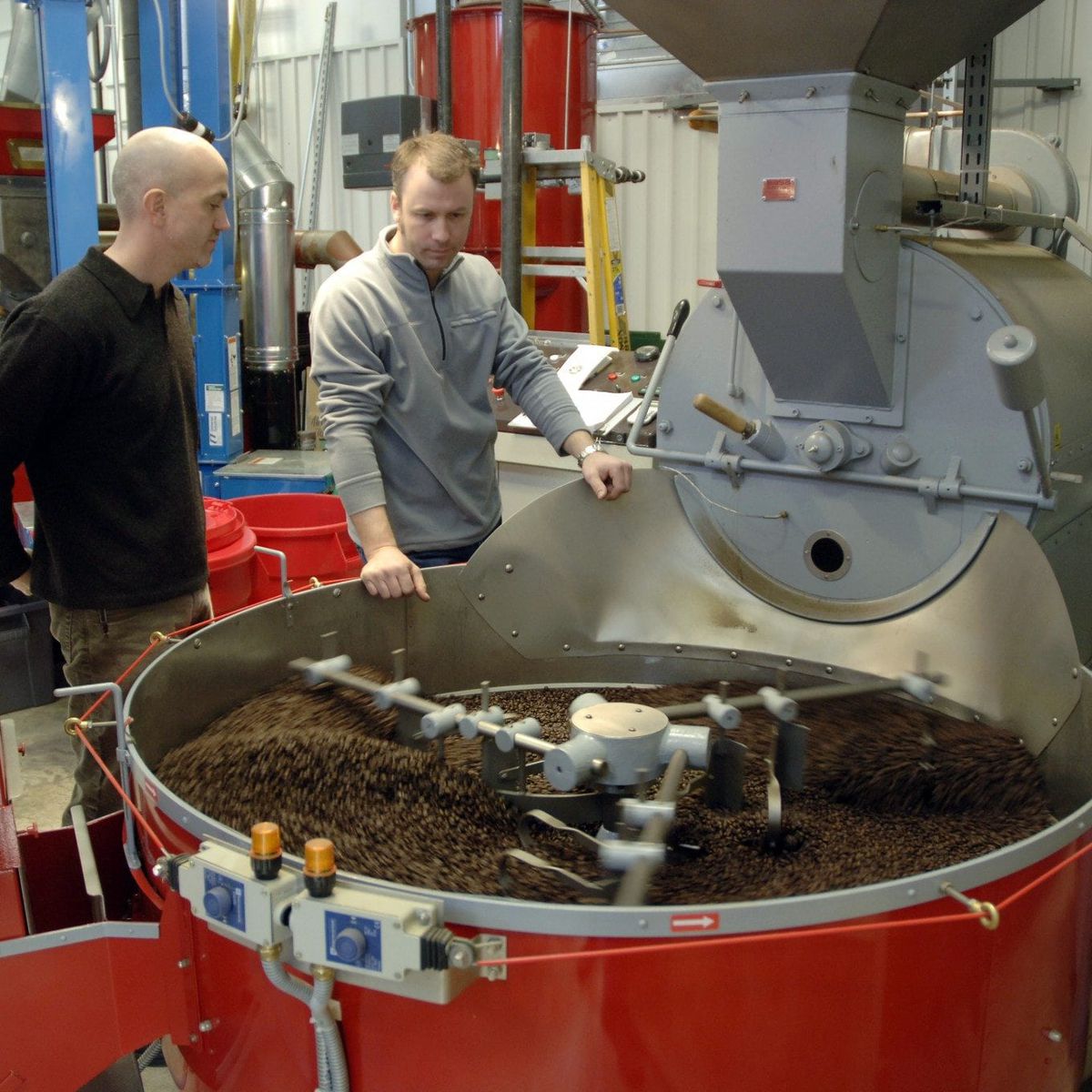 Two men stand in a coffee roastery, looking down into a piece of equipment full of coffee beans