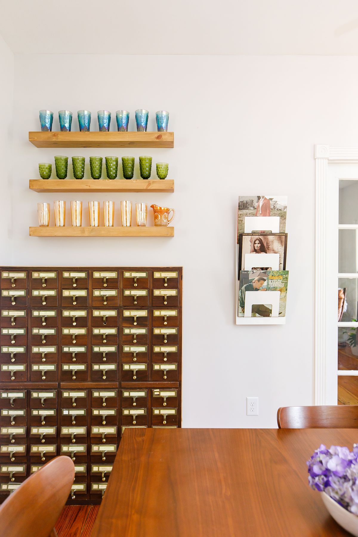 A card catalog sits against a white wall with shelves that hold multiple colored drinking glasses and a magazine rack full of magazines. There is a table in the foreground.