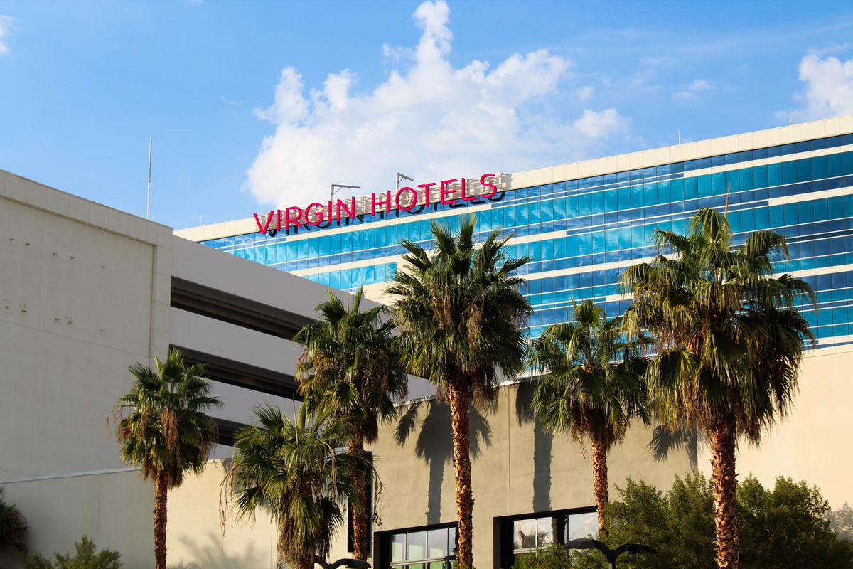 The exterior of a hotel with a red sign on top, blue windows, palm trees, and a parking garage