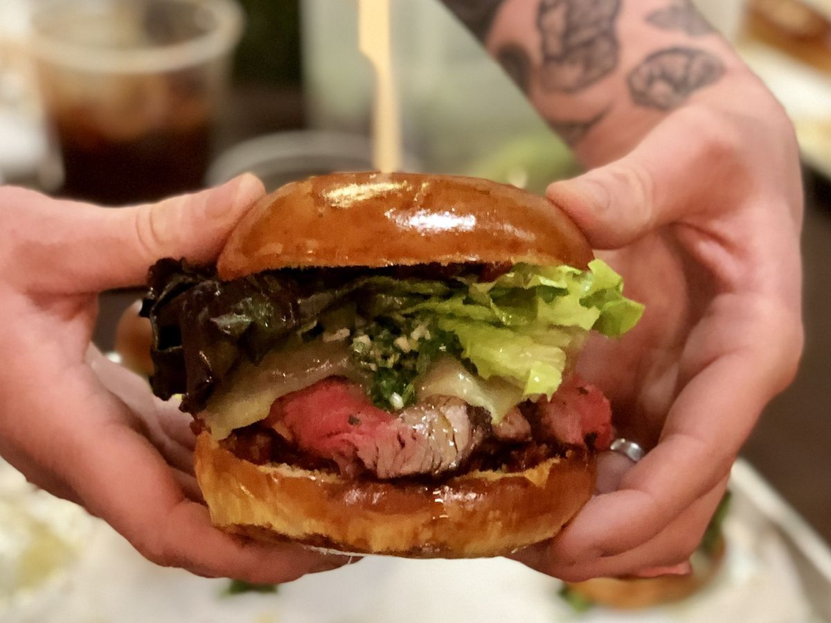 Two hands holding a slow-roasted tri-tip, spiced tomato jam, and chimichurri sandwich on a bun.