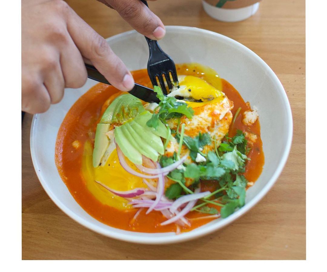 Two hands holding a knife and fork cut into huevos rancheros in a white bowl