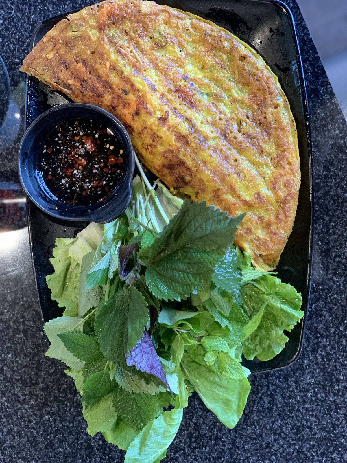 An overhead shot of a crisp egg pancake on a black plate with greens on the side.