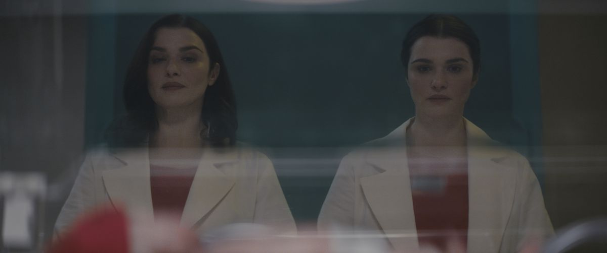 Two identical women (Rachel Weisz) stand side by side one another in matching white doctor’s outfits staring at a baby in a hospital bassinet.