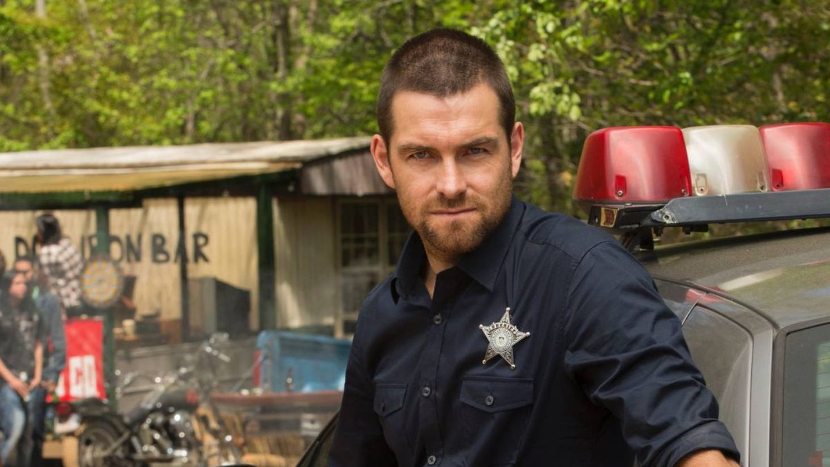 Antony Starr, with a shaved head, wears a police officer’s uniform and leans against a cop car in Banshee.
