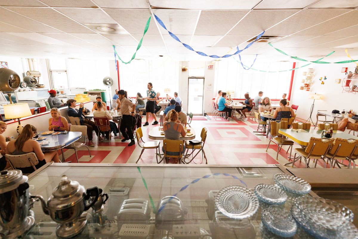 A view from the order counter into a retro dining room filled with natural light, and colorful streamers hanging from the ceiling as people dine on a Saturday morning.