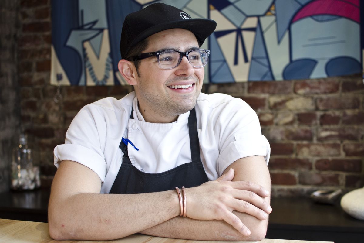 Portrait of a chef wearing glasses, a black apron, a white shirt, and a black baseball cap. He sits in front of a brick wall, arms crossed, looking off to the side and smiling.