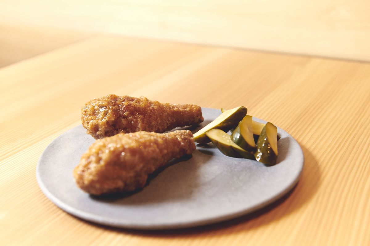 Two of Momofuku Ko Bar’s chilled fried chicken drumsticks sit on a ceramic plate, adjacent a piles of pickles