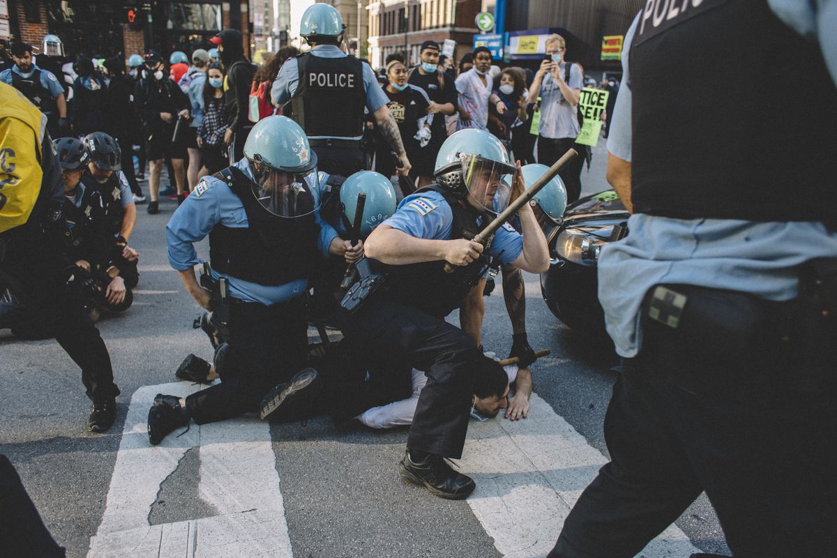 Four officers in light blue shirts, navy slacks, and light blue riot helmets pin down a man in the middle of the street. One officer is getting to his feet, and has a wooden baton raised as if to strike. Others are pinned to the ground by officers in the background.
