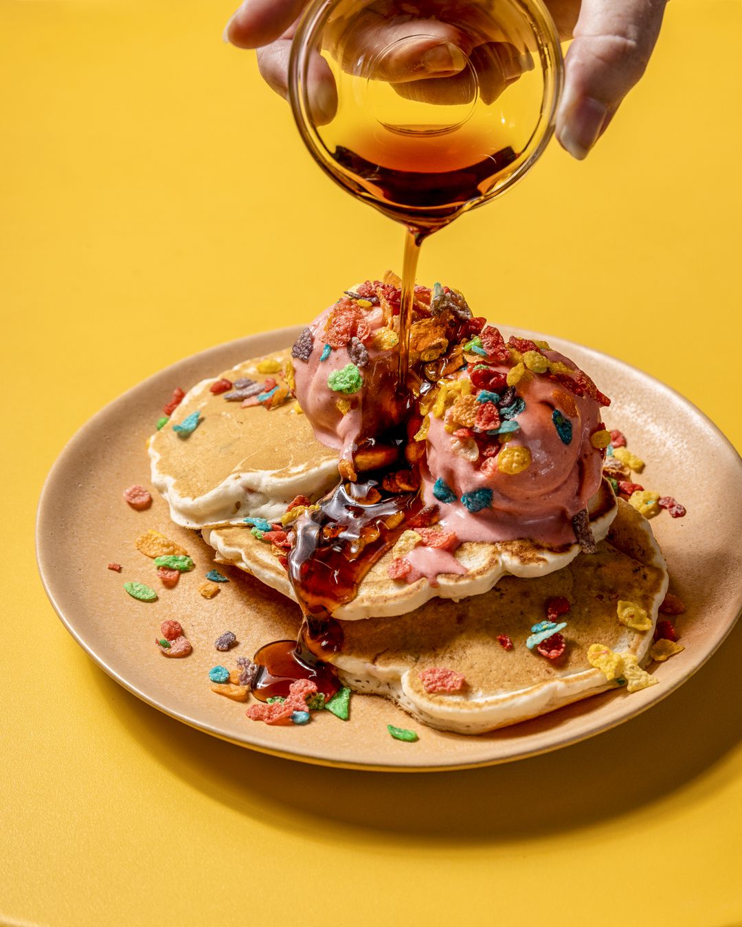 Three pancakes with cereal sprinkles, two scoops of pink ice cream, and syrup being poured from a clear glass cup by a hand