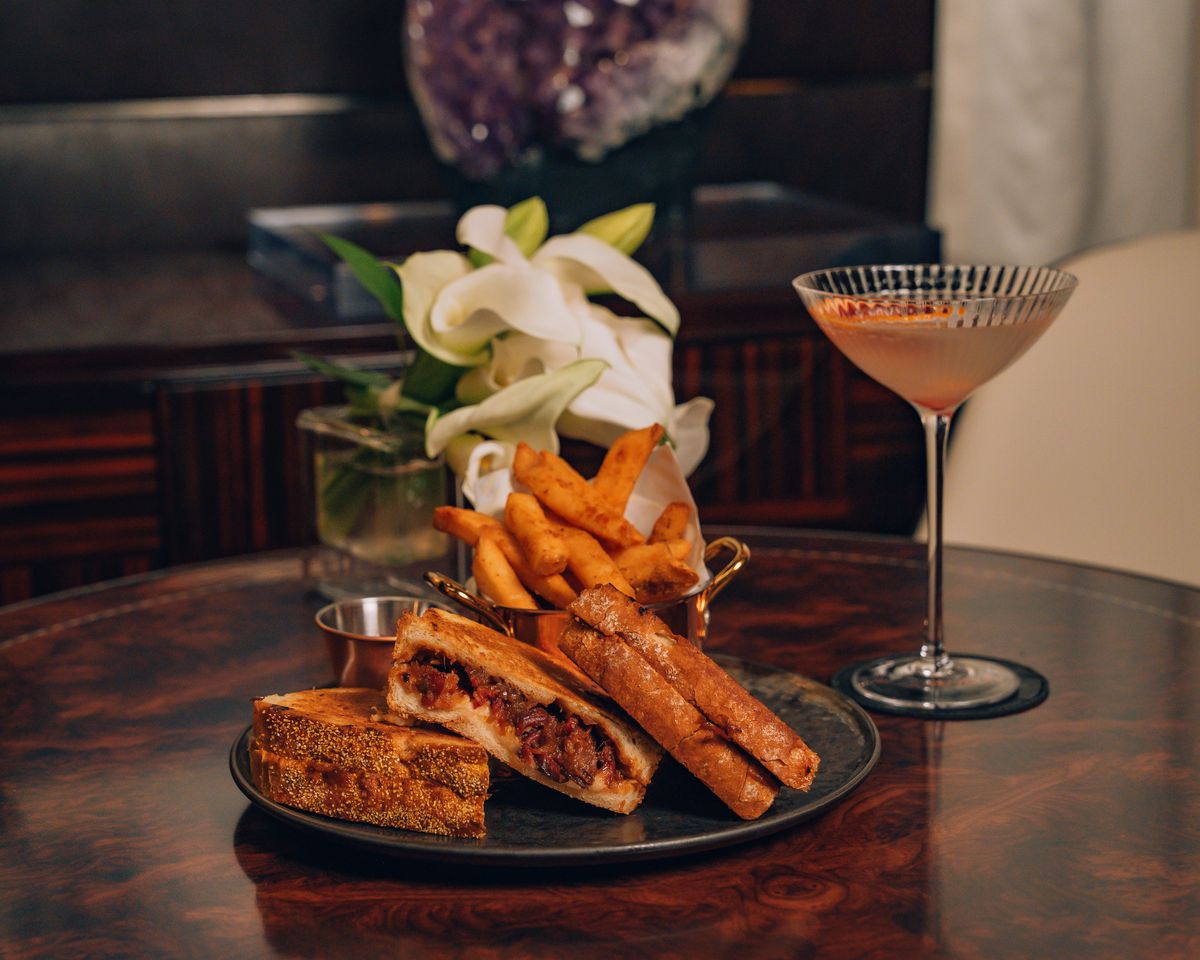 A grilled cheese sandwich cut into thirds and stacked on a plate, with a cup of fries and a cocktail in the background.