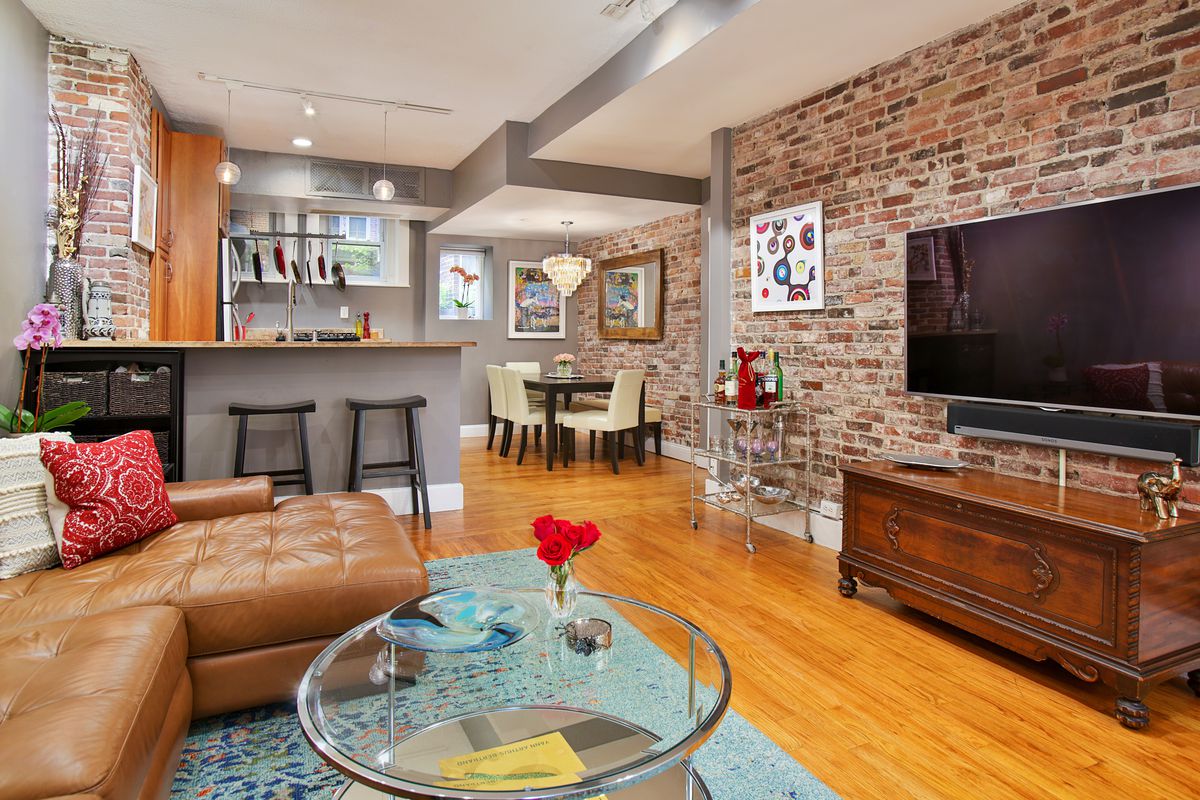 A living room with a large couch facing a TV, and there’s a lot of exposed brick on the walls, and the living room leads to a kitchen. 
