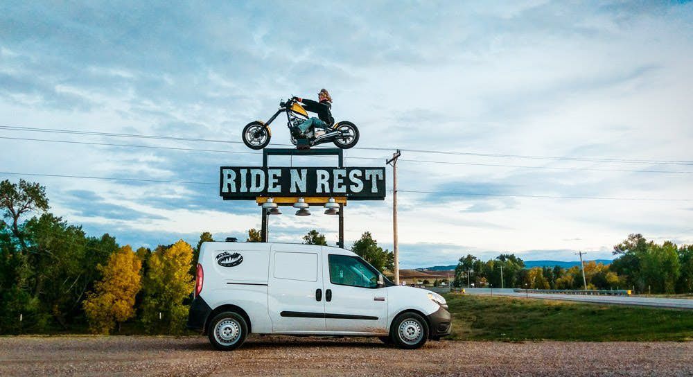 A white camper van parking in a clearing. In the background is a sign that has words that say: Ride N Rest. 