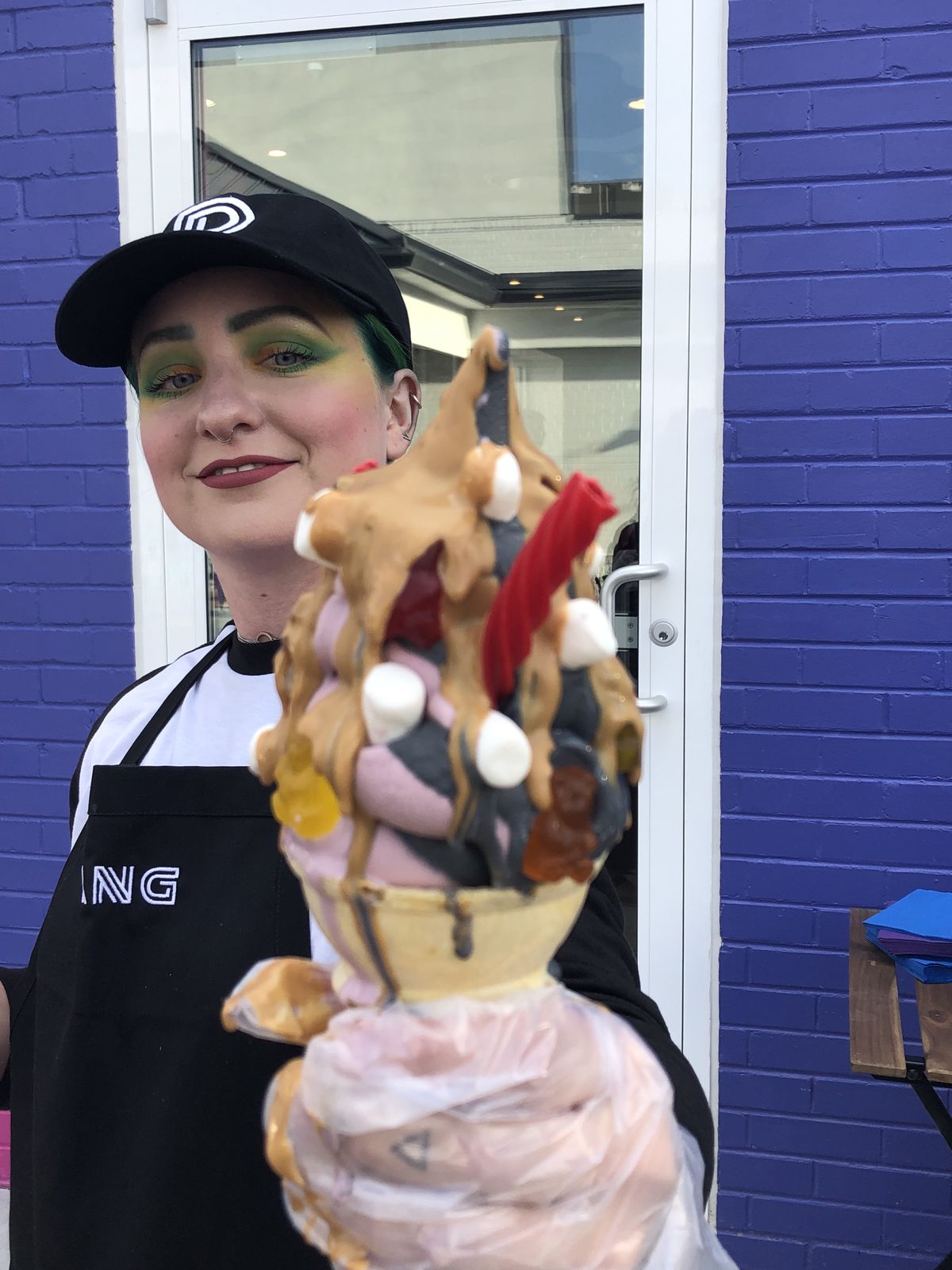 A photo of a manager at Dang holding up a cone of swirled purple and gray soft serve adorned with cookie butter sauce, marshmallows and licorice 
