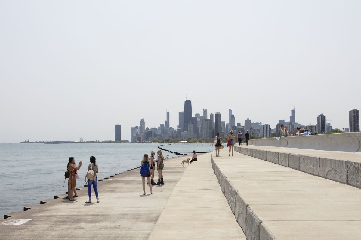 A tiered concrete breakwater curves around the edge of the a lake with small groups of people strolling in the sun with tall towers and buildings in the distance.