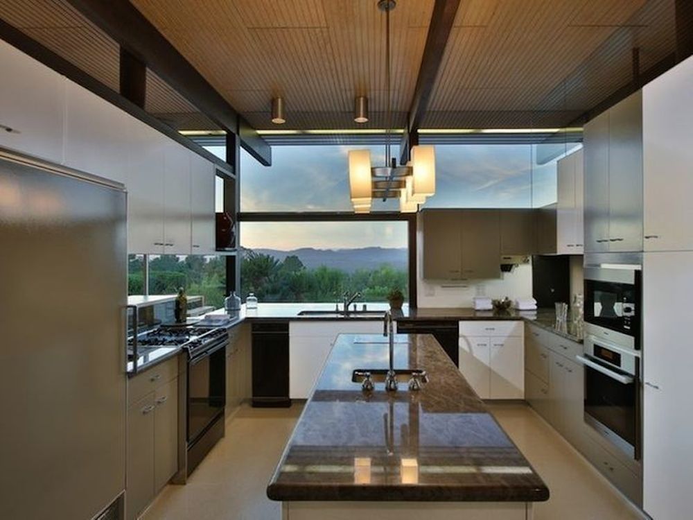 A midcentury modern kitchen with silver and white cabinetry and a wooden ceiling. There is a kitchen island with a granite countertop. There are windows overlooking a view with trees and mountains.