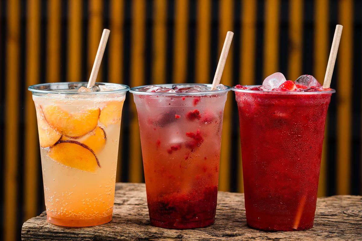Three plastic cups filled with red and orange liquid, slices of fruit, and straws, are lined up on an outdoor table.