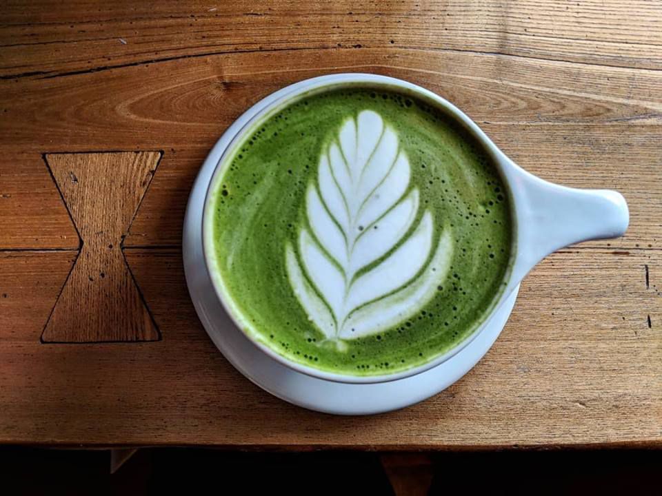 Overhead shot of a bright green matcha latte with fern foam art. It’s in a white mug, sitting on a wooden counter top.