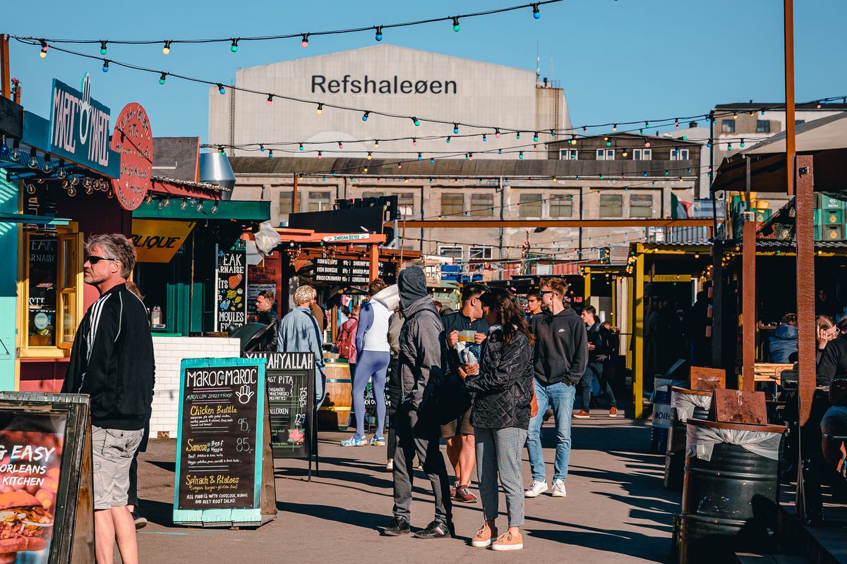 A crowd at outdoor market stalls, with a large building beyond printed with the word Refshaleoen.