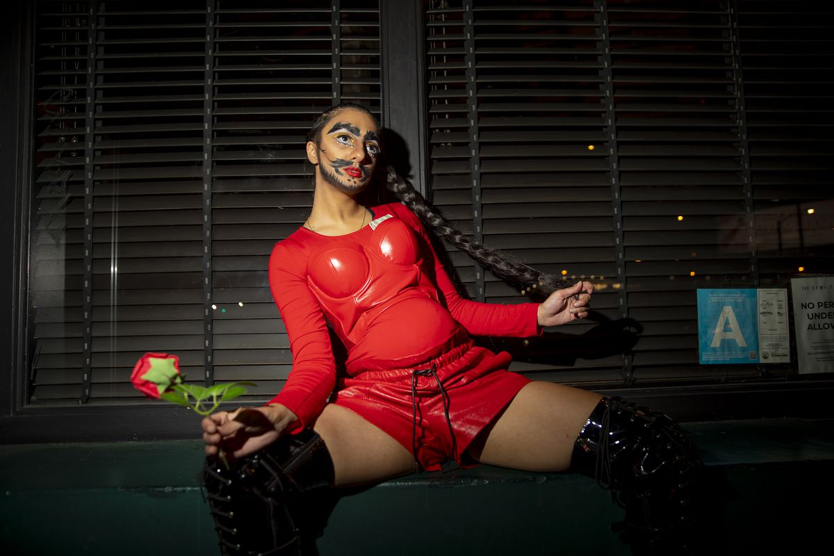 Posing, legs spread, in red bodysuit and fake belly, rose in one hand and long braid in another, bills tucked into bustier.