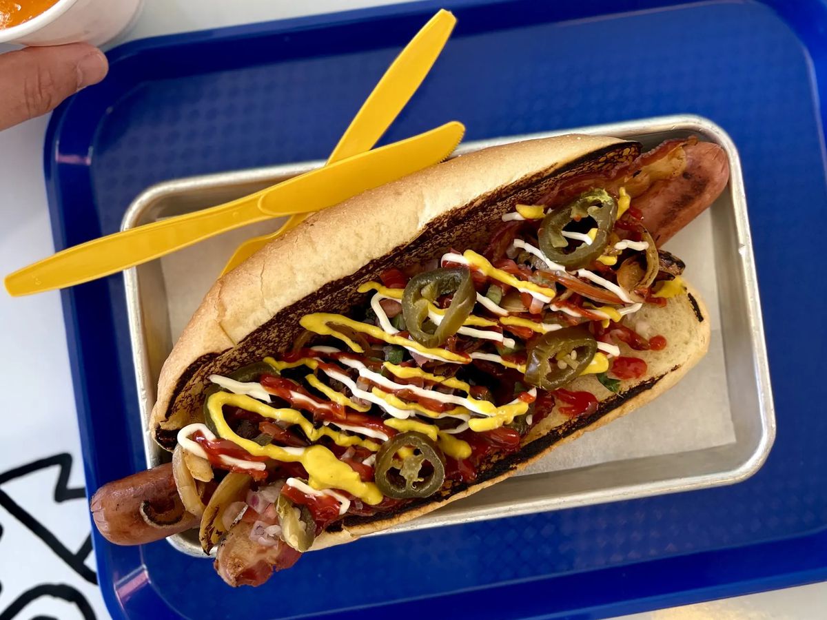 A foot-long hot dog toppped with jalapenos and drizzled with cheese sitting on a blue tray with yellow utensils on the side