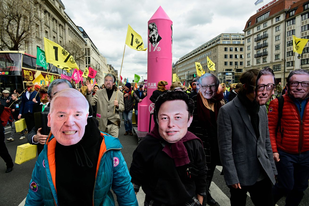 Protesters march with masks of billionaire faces, with a mask of Elon Musk in the center.