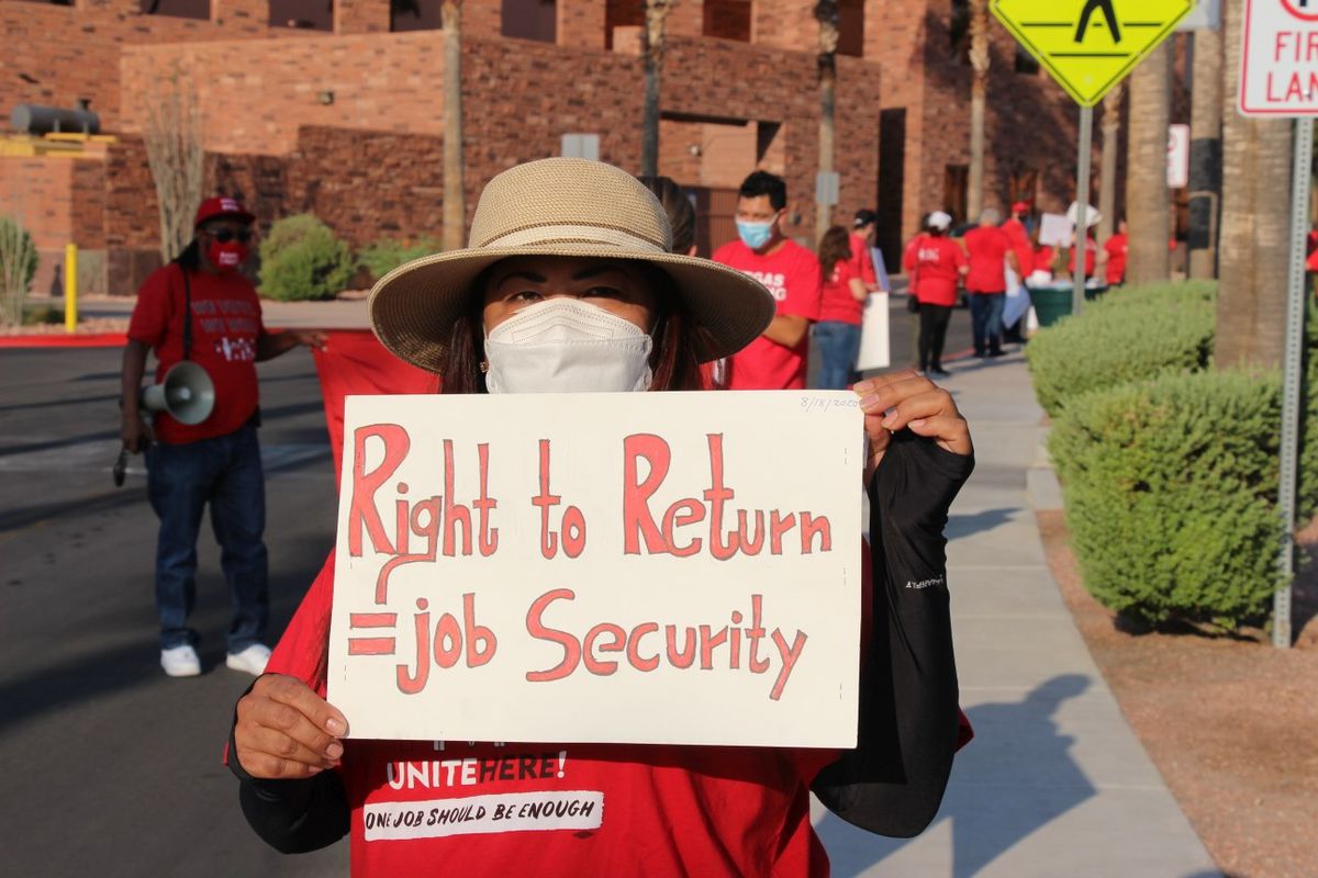 A person in a red shirt, hat and mask holds a sign that says Right to Return = Job Security outside with a line of people in red T-shirt behind