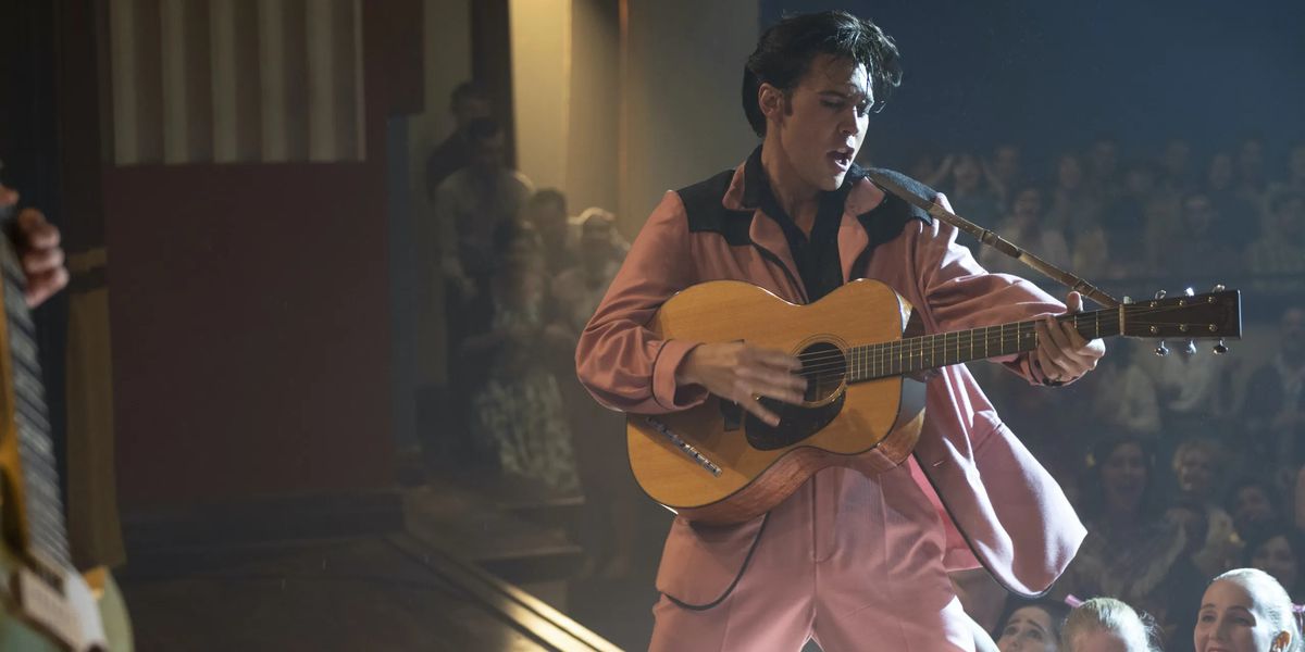 Em um smoking rosa e preto, Elvis toca guitarra no palco.