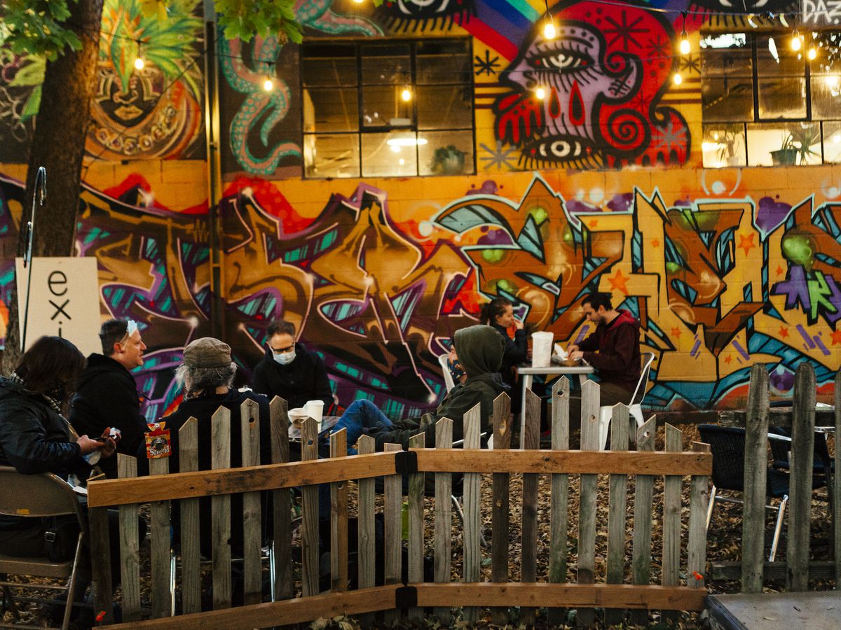 In the foreground is a wooden fence; behind it are diners dressed in warm clothing, and a bright, colorful graffiti mural made with mostly orange, blue, and red colors, lit by globe lights in an outdoor space. 