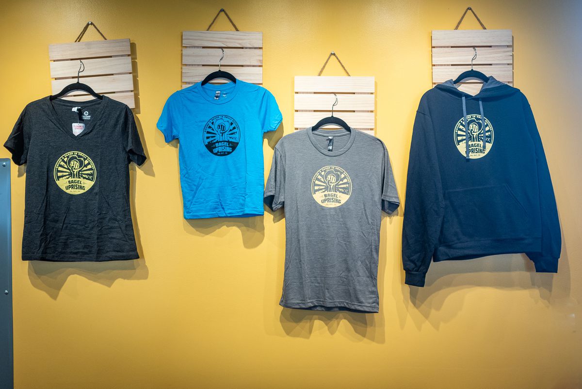 T-shirts and hoodies with the Bagel Uprising logo hang on a display wall