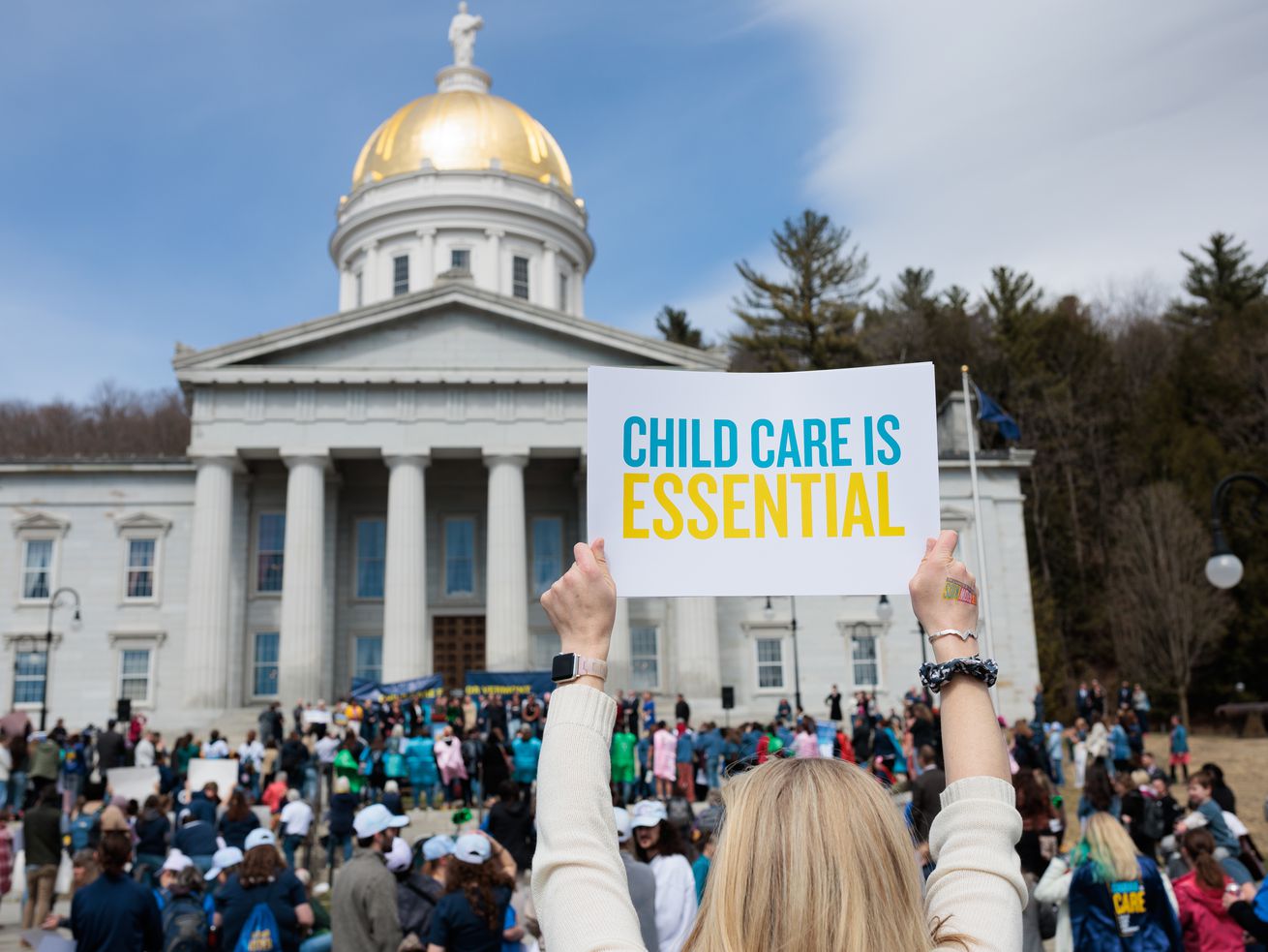 A protester in a crowd in front of a domed government building holds a sign that reads “Child care is essential.”
