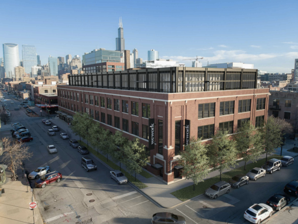 An aerial view of a long brick vintage warehouse buildings with a more contemporary dark metal fourth floor addition. Taller buildings are visible on the horizon. 