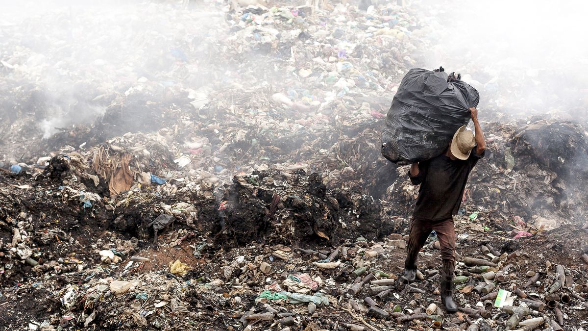 A person carrying a bag of garbage across a steaming garbage dump.