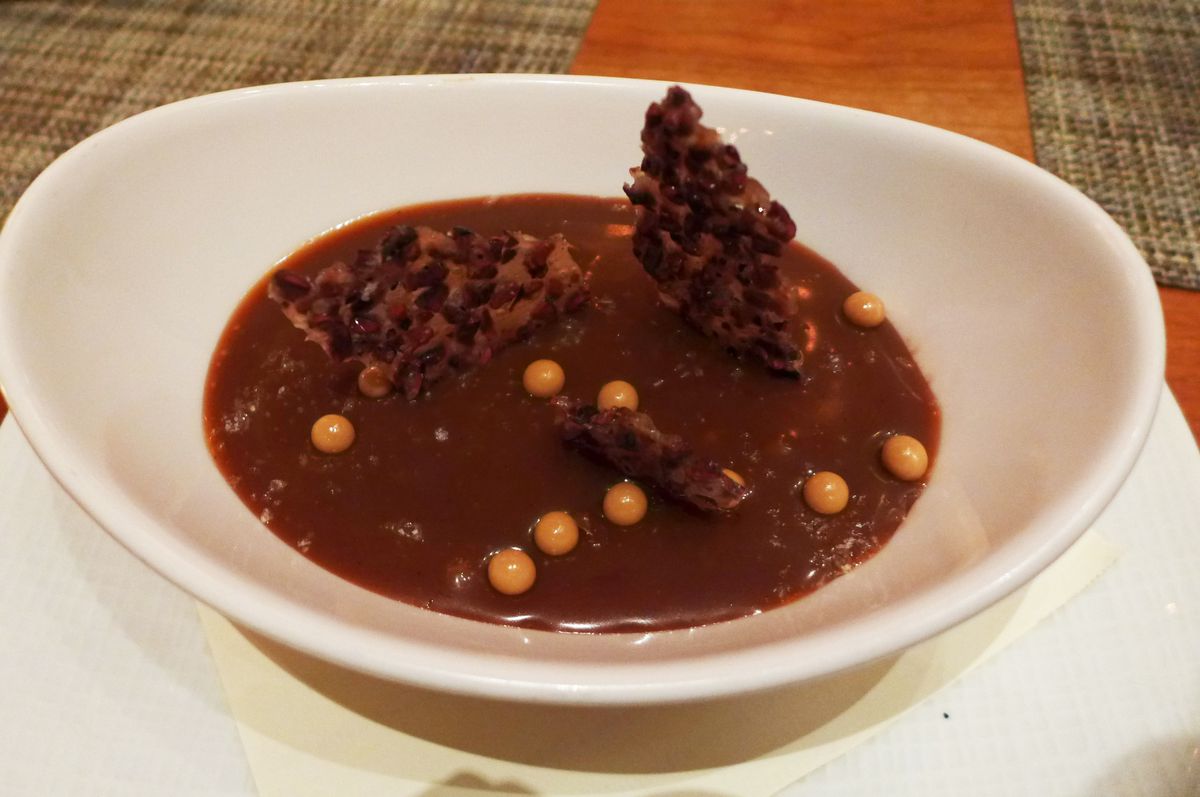 Chocolate pudding in a white bowl with brown beads of salted caramel and a chocolate bark sticking out of the top.