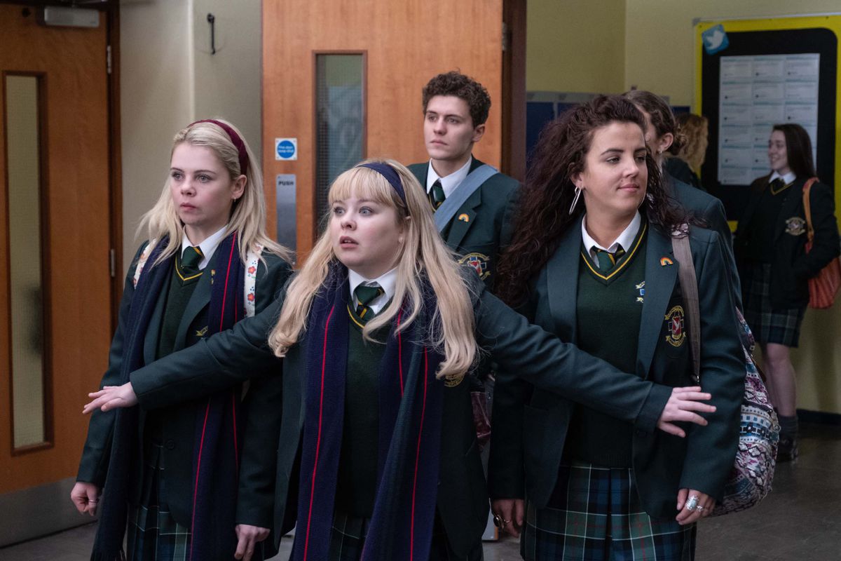 The Derry Girls wear their school uniforms. One stands in front of the rest with her arms outstretched.