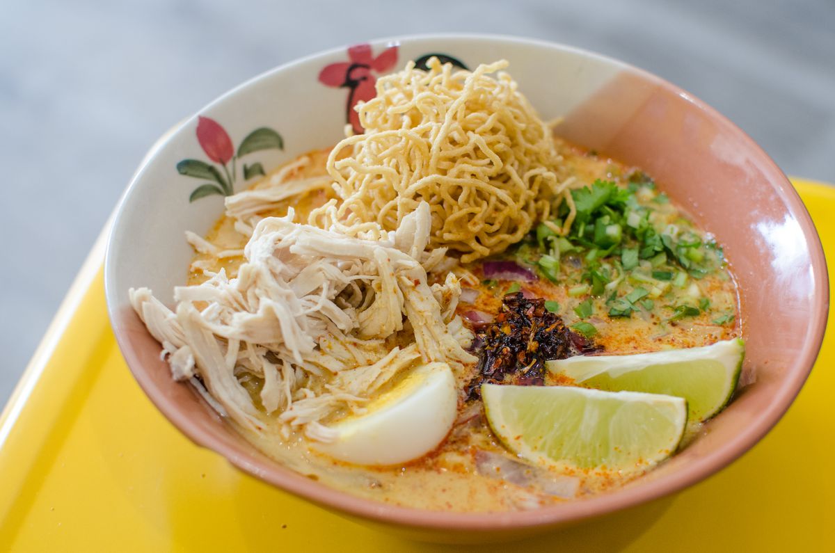 Khao soi —&nbsp;a yellow curry with chicken, egg, lime, crispy noodles, and more —&nbsp;is served in a traditional Thai-style bowl decorated with a rooster. The bowl sits on a yellow surface.