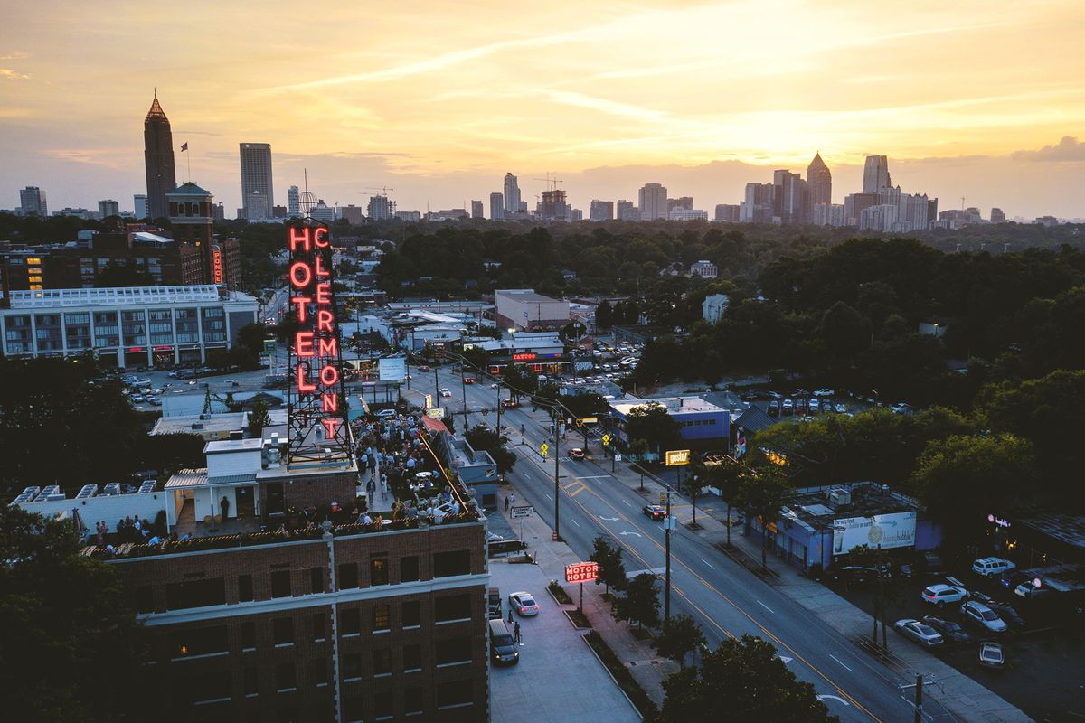 An aerial overlooking the rooftop filled with people at the bar of the Hotel Clermont on dusk with the Atlanta skyline backlit by the setting sun in the distance