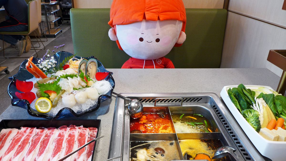 A stuffed tomato character with a red hat sits opposite me at a booth, with a hot pot meal spread before us