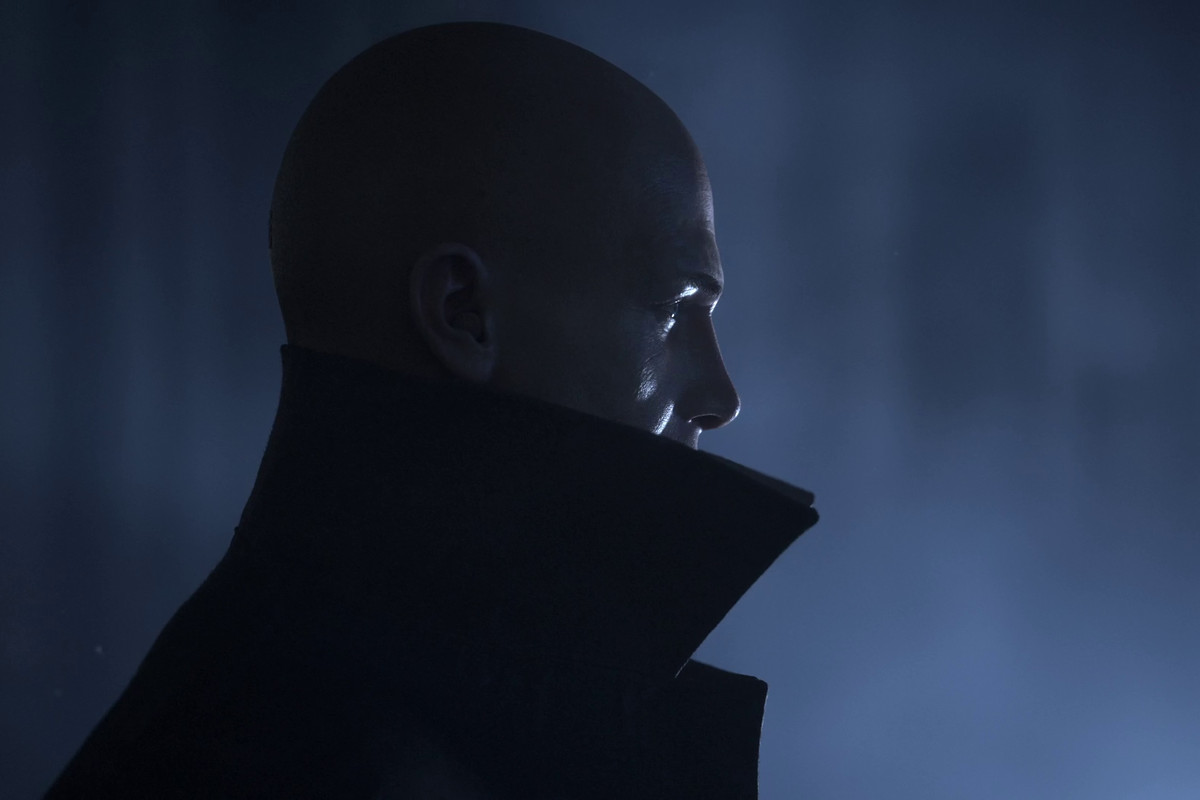 Hitman 3 - Agent 47 in profile, wearing a high collared coat. He’s backed by an ominous looking blue light.