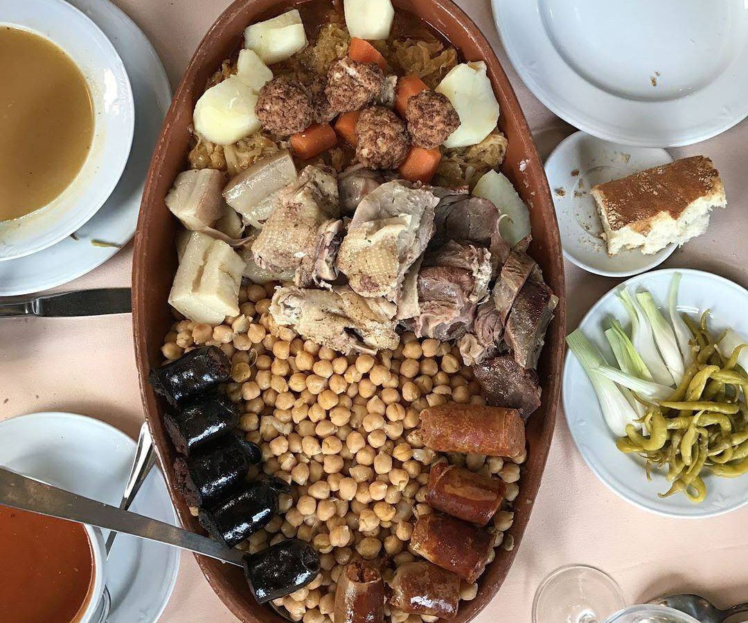 A large tray with different cuts of meat and chickpeas in the center of several other half-eaten dishes, including vegetables, breads, and soups.