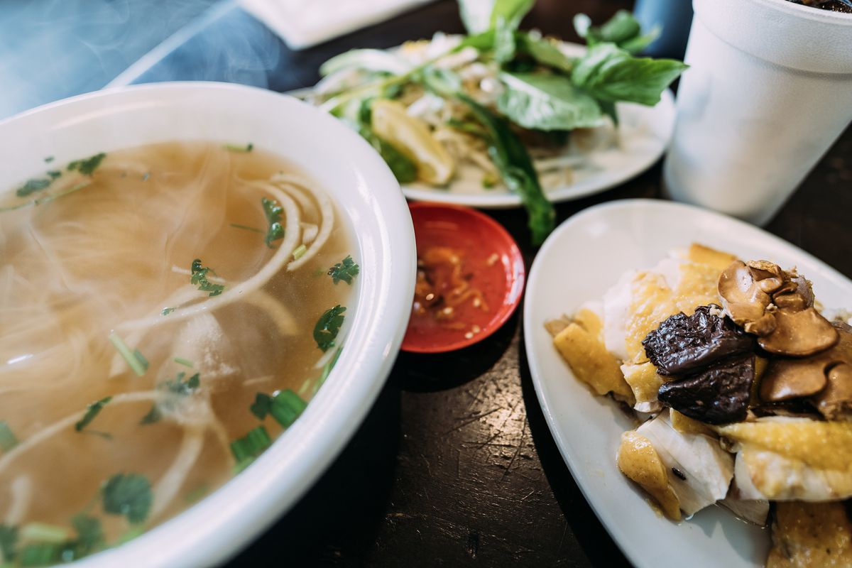 A bowl of pho and a plate of meat on the side with herbs and spices behind it.