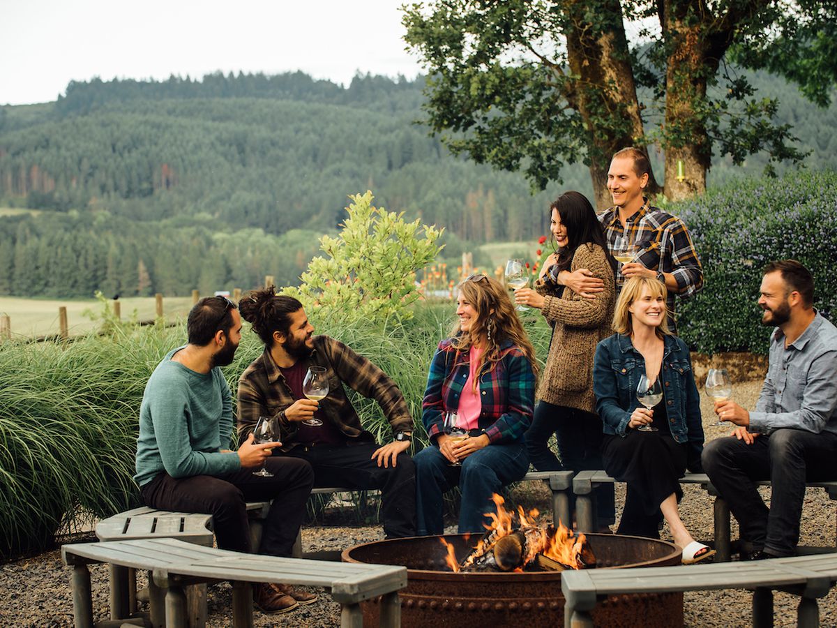 A group of 20somethings and 30 somethings sit around an outdoor firepit with glasses of wine, surrounded by fields and farmland.