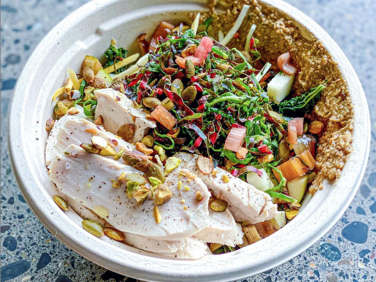 A beige bowl is filled to the top with greens, veggies, poached chicken, and pumpkin seeds.