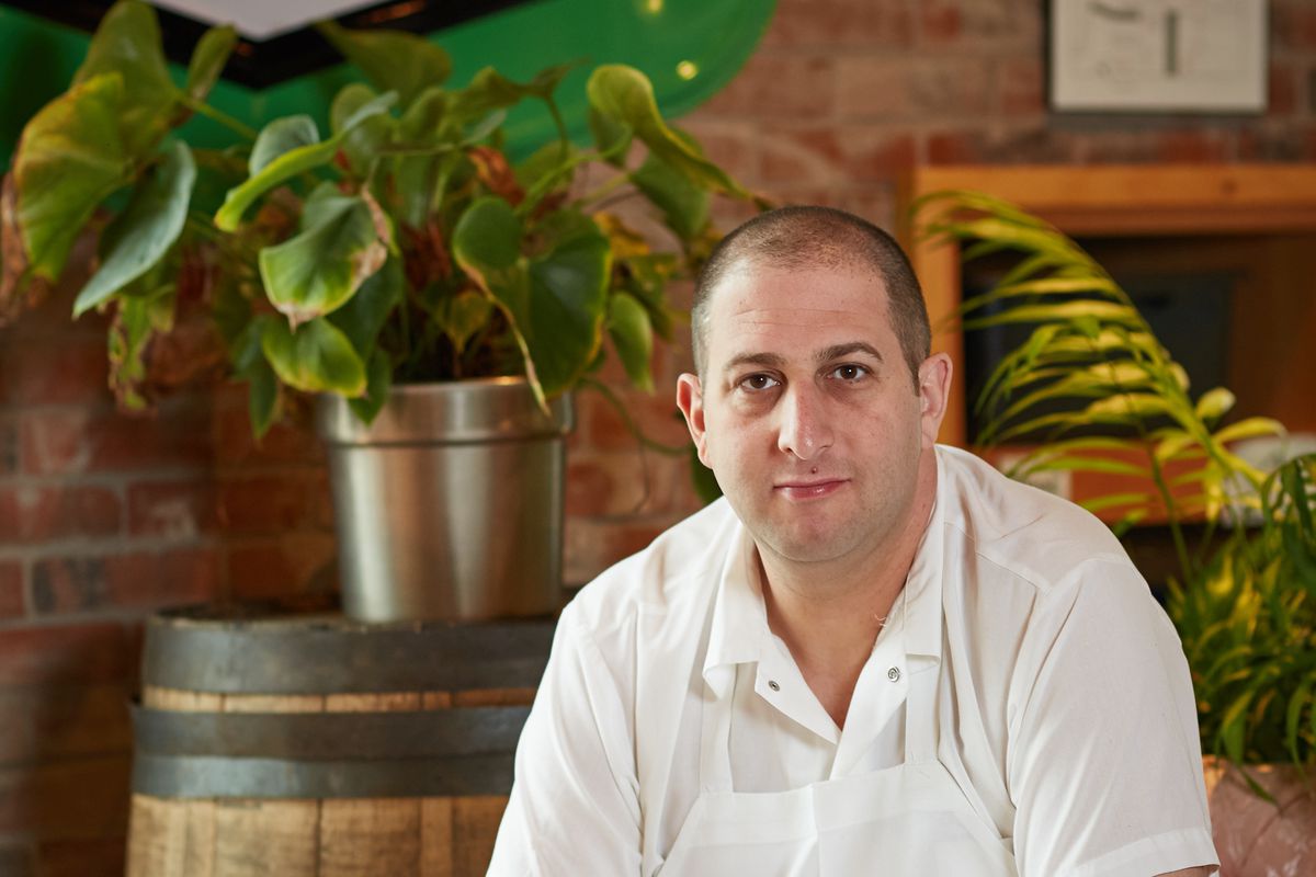 A photo of chef Steve “Nookie” Postal, wearing chef’s whites and sitting in front of a barrel and greenery in a restaurant