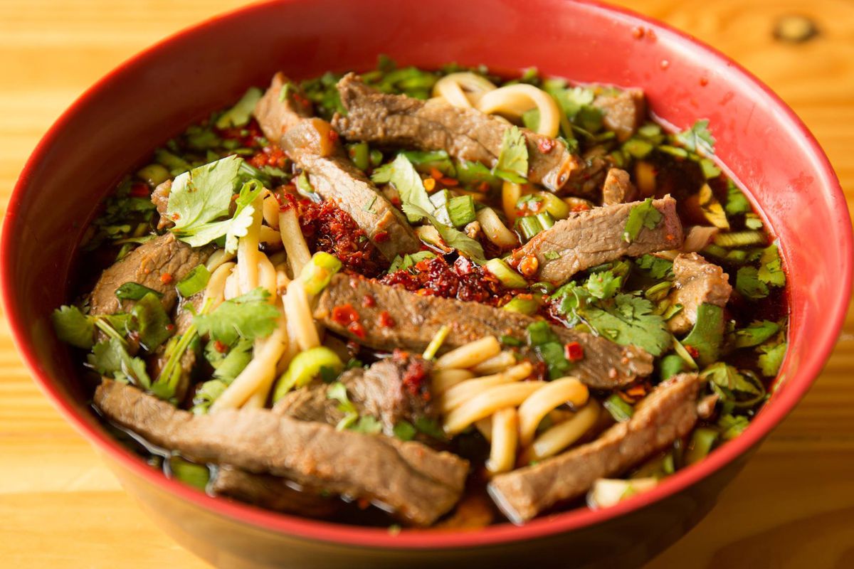 Noodles and beef floating in a spicy broth in a red bowl. The bowl is garnished with chile flake and cilantro leaves.