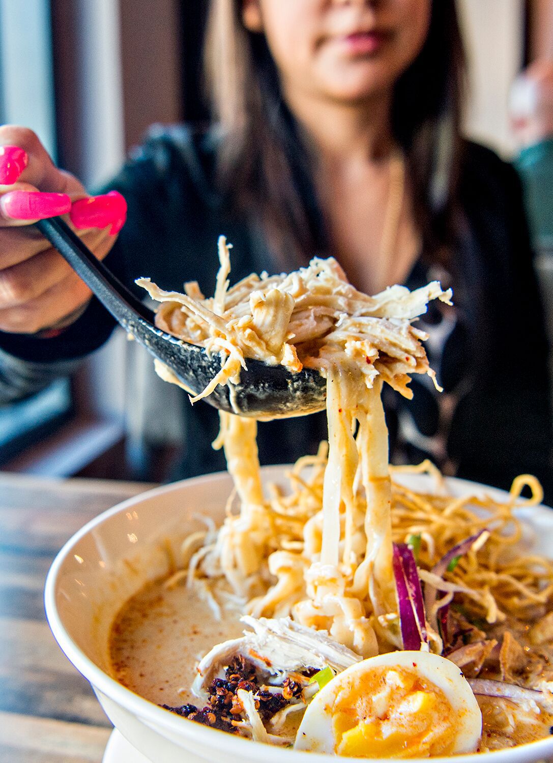 A photo of a woman reaching for spoonful a of the chicken and noodles in the Khao Soi Kai